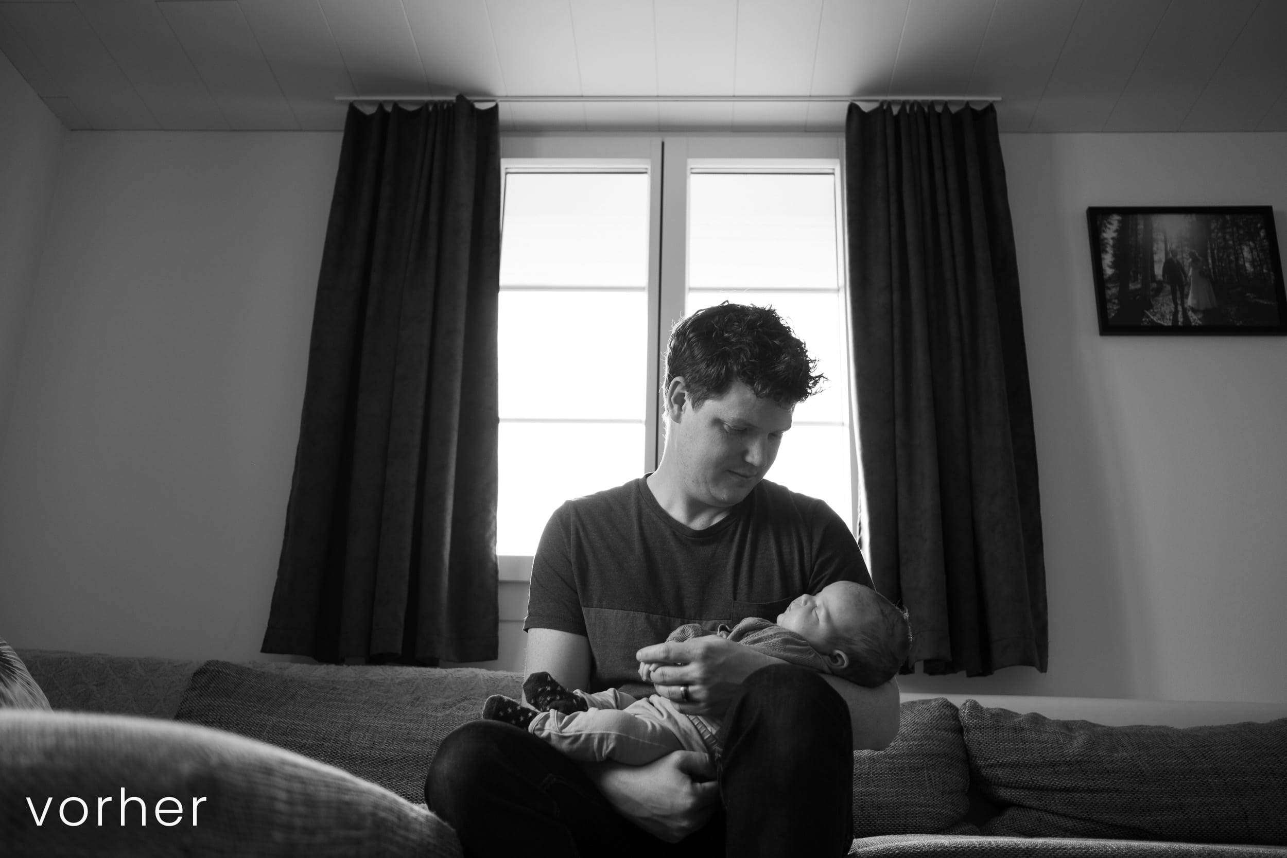 Vater sitzt mit Baby auf Sofa vor Fenster.